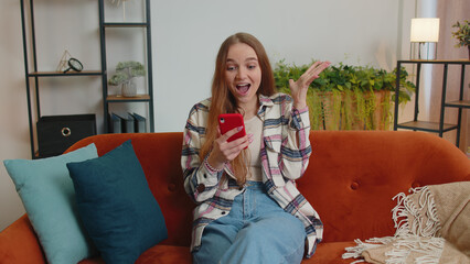 Excited happy joyful stylish teen girl in sweater use mobile cell phone typing browsing say Wow yes found out great big win news doing winner gesture. Young adult woman at home in living room on sofa