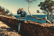 © skyNext - A spruce black guy in a stylish blue costume is holding his straw hat from the wind while sitting on a stone fence outdoors next to the ocean coast surrounded by trees on a warm sunny day