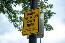 Low Angle View Of A Park At Your Own Risk Sign Outside Of A Baseball Field, Warning Anyone Parking Their Cars Of The Chance Of Fly Balls