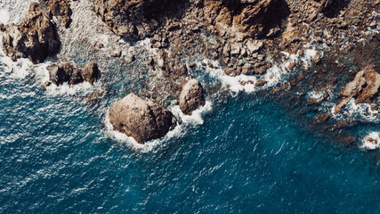 Aerial top view of waves break on rocks in a blue ocean. Sea waves on beautiful beach aerial view drone 4k shot. Bird's eye view of ocean waves crashing against an empty stone rock cliff from above.