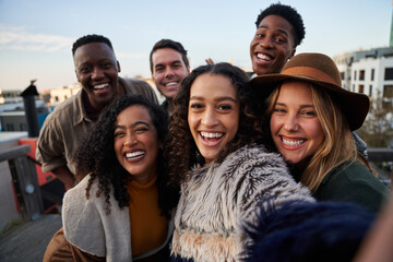 Multi-cultural group of friends taking a selfie at a rooftop party. Close up, smiles with diverse young adults.