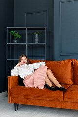 Sticker - Portrait of fashionable young woman in a beige skirt, white blouse and stylish beige black seude shoes with a buckle posing on a orange sofa.