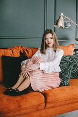 Sticker - Portrait of fashionable young woman in a beige skirt, white blouse and stylish beige black seude shoes with a buckle posing on a orange sofa.