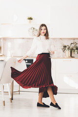 Wall Mural - Portrait of fashionable woman in a red skirt, white blouse and stylish suede shoes with a buckle posing on the kitchen. Girl with a vine glass in a hands