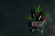 Supermarket trolley with marijuana leaves and medical cannabis oil bottle on black background. Copy space.