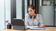 © amnaj - Charming young Asian businesswoman sitting smiling working with document and laptop on the desk in the office.