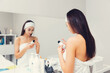 © _KUBE_ - A young woman in a bath towel with a bandage on her head, holding a bottle of cosmetics and reading the composition of the product, standing in the bathroom. The concept of home cosmetic skin care