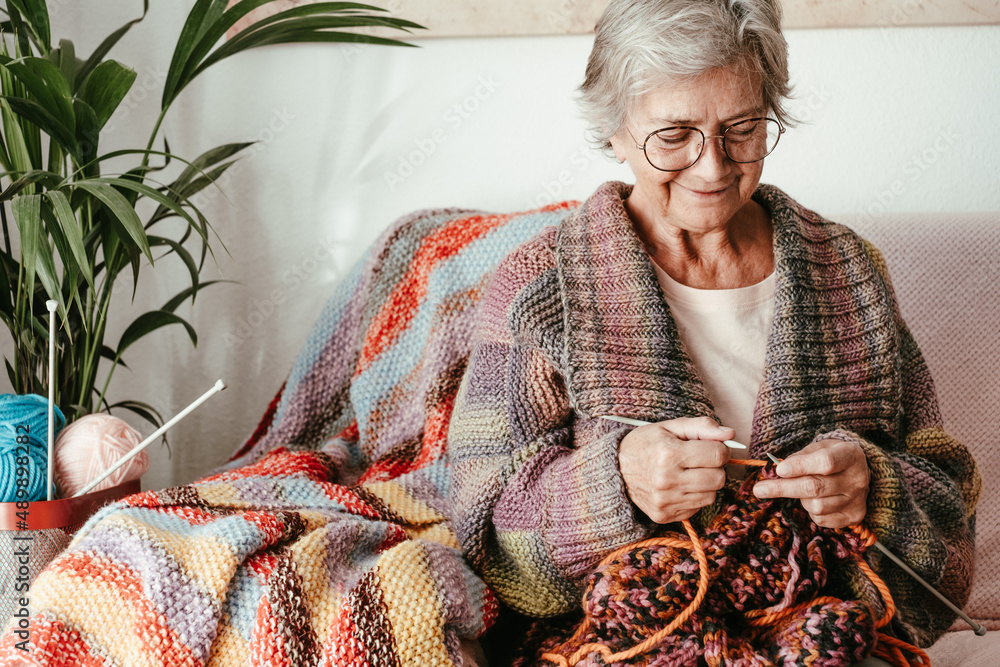 Senior mature woman sitting on sofa at home while knitting and looking ...