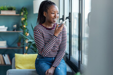 Smiling Young African American Woman Sending Audio Message With Mobile Phone While Sitting On Sofa At Home