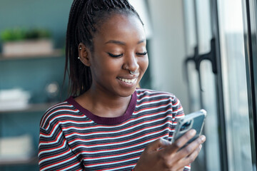 Wall Mural - Smiling young african american woman using her mobile phone while standing near the window in living room at home.