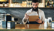 © Tijana - Handsome Barista Doing an Inventory of the Products in a Coffee Shop. Serious waiter with a beard standing in the bar counter and using clipboard for counting stock in the restaurant.