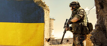 Ukrainian Soldier Military In The War With A Weapon In His Hands. The Flag Of Ukraine Is Painted On A Brick Wall. Relations Between Ukraine And Russia