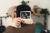 Fototapeta Mapy - Happy couple hugging, caucasian woman holding ultrasound scan of her baby, focus on foreground. High quality photo