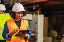 Engineer Asian And African Woman Wearing Safety Helmet And Vest Holding Clipboard And Take Note On The Paper In The Automotive Part Warehouse.Products And Corrugated Cardboard.  