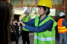 Female Warehouse Worker In Safety Uniform And Hard Hat Works By Hydraulic Jack Lift Piles Of Cardboard For Shipping And Logistic Transport At Manufacture Factory, Supply Packaging Stocks Industrial.