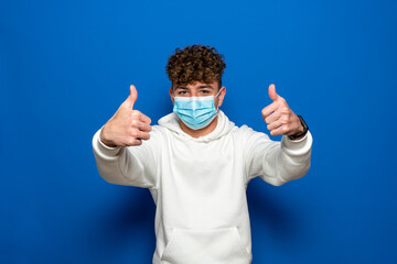 Attractive man with curly hair wearing a surgical mask to protect himself from coronavirus with thumbs up isolated on blue studio background. Pandemic concept