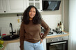 © Anatoliy Karlyuk - Beautiful 20-years-old African American girl in brown jumper and blue jeans standing against lovely cozy white kitchen, with hand on her waist, smiling, having long black curly hair