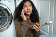 © Anatoliy Karlyuk - Close-up picture of pretty good-looking dark-skinned woman holding tools in hands and calling out appliance service to repair her broken washing machine, sitting on kitchen floor, looking worried