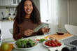 © Anatoliy Karlyuk - Beautiful woman of 20s cooking breakfast, cutting avocado and other vegetables, tomatoes and spinach on table in front of laptop, pretty female smiling, being in perfect mood on Monday morning