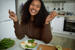 © Anatoliy Karlyuk - Picture of cheerful happy woman with dark skin and long curly hair sitting at table in front of fried eggs decorated with spinach leafs and avocado slices. Healthy food and lifestyle