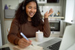 © Anatoliy Karlyuk - Young adorable sweet woman of black ethnicity studying online, joining video conference with her group mates, smiling, waving, sitting at kitchen table in front of laptop, making notes of lecture