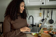 © Anatoliy Karlyuk - Pretty happy dark-skinned female in brown clothes holding laptop, videochatting with her friend while cooking breakfast in late morning of lazy weekend, standing next to table top with vegetables