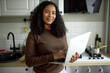 © Anatoliy Karlyuk - Beautiful charming young lady in casual brown jumper holding laptop in hands standing at kitchen with back turned to table top and oven, searching recipe to cook something delicious for dinner