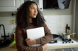© Anatoliy Karlyuk - Cute adorable romantic-looking thoughtful young girl standing against kitchen furniture, looking through window holding closed laptop in hands after doing homework, having beautiful black curly hair