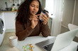 Charming black ethnicity female of 30s sitting at kitchen table in front of opened laptop, cup and vitamin pills, holding bottle of food supplements, reading suggested usage and warnings
