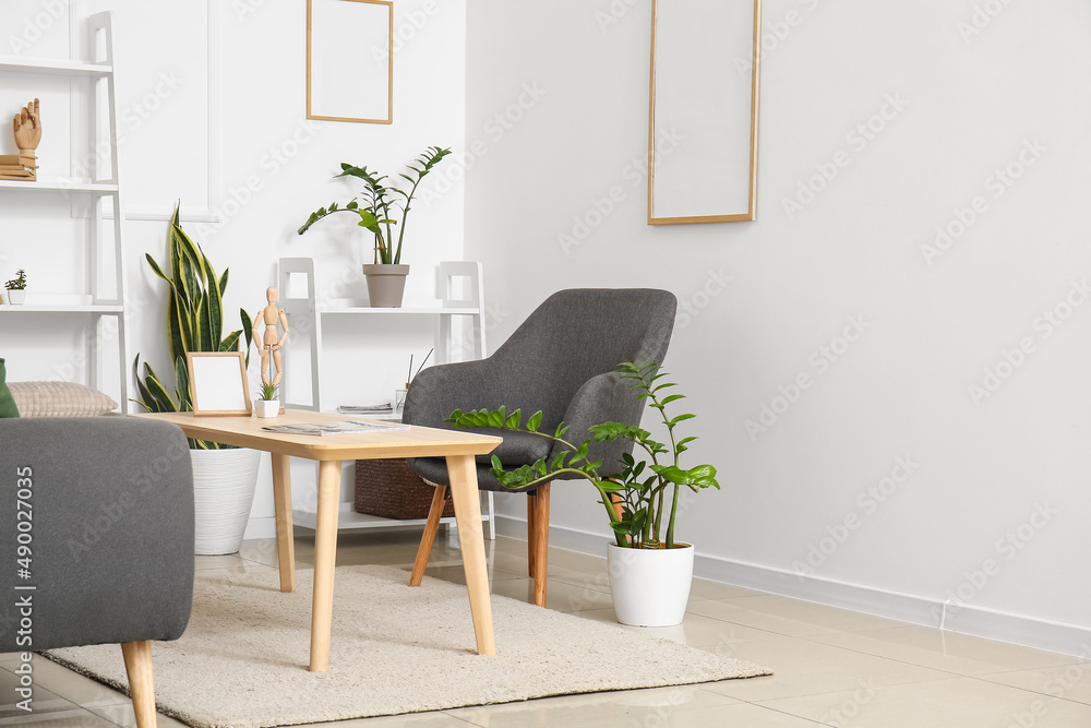 Comfortable armchair, table, houseplants and shelving unit near white wall