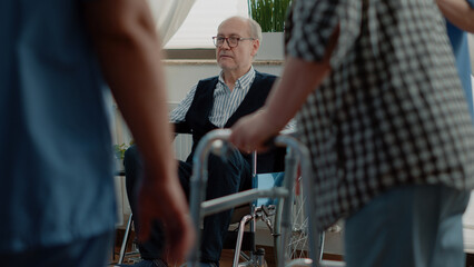 Old disabled man relaxing in nursting home room. Retired person with disability sitting in wheelchair preparing for recovery and wellness while nurses helping patient with walk frame