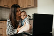 © Ananass - a young mother with many children working on a laptop sitting in the kitchen with a baby in her arms while the eldest daughter helps with cooking and cleaning in the kitchen