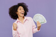 © ViDi Studio - Young smiling happy fun woman of African American ethnicity 20s in pink striped shirt holding fan of cash money in dollar banknotes do winner gesture isolated on plain pastel light purple background