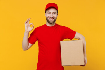 Wall Mural - Professional delivery boy employee man 20s in red cap T-shirt uniform workwear work as dealer courier hold face with cardboard box show ok gesture isolated on plain yellow background. Service concept.