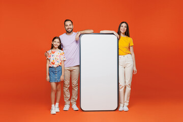 Wall Mural - Full body young parents mom dad with child kid daughter teen girl in basic t-shirts big blank screen mobile cell phone with copy space mockup area isolated on yellow background. Family day concept