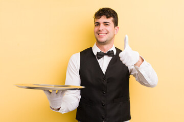Wall Mural - young handsome man feeling proud,smiling positively with thumbs up. waiter and tray concept