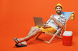 Young tourist man wear beach shirt hat lie on deckchair hold use work on laptop pc computer show thumb up isolated on plain orange background studio portrait. Summer vacation sea rest sun tan concept