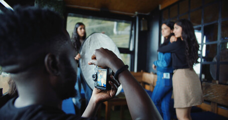 Wall Mural - Back shot of a black male holding a camera shooting on posing blurred South Asian girls indoors