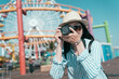 © PR Image Factory - Smiling woman in sunglasses and hat stand with professional camera in hand and happily looking lens while spending time in amusement park with ferris wheel on background. female travel photographer