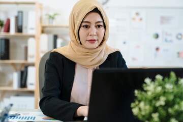 Wall Mural - Arabian Businesswoman working in workplace. young muslim office lady with hijab concentrated typing on keyboard laptop computer. confident islam female employee in black suit do project on notebook