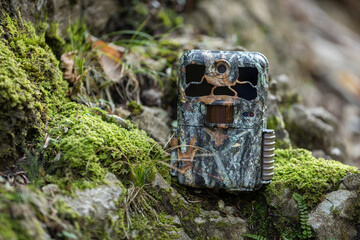 Camouflaged trail camera standing on a moss covered rocks in nature. Wildlife monitoring device with and infrared illumination and PIR motion sensor hidden in forest.