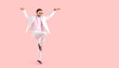 Funny man posing in fashion studio. Full body happy attractive young guy wearing white suit, pink shirt, cool sunglasses and trainers standing on one foot, arms spread apart, on text space background