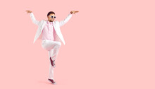 Funny Man Posing In Fashion Studio. Full Body Happy Attractive Young Guy Wearing White Suit, Pink Shirt, Cool Sunglasses And Trainers Standing On One Foot, Arms Spread Apart, On Text Space Background