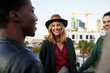© StratfordProductions - Caucasian female smiling with group of multi-cultural friends socializing on a rooftop terrace at dusk.
