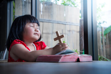 Wall Mural - Little girl praying and holding wooden cross on bible at home, child's pure faith