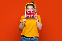 Learning British English, Student Exchange And Travel. Happy Lady Holding Small Flag Of UK, Looks At Camera. Girl Peeking Out From Behind Flag. Ready To Study In College, Standing On Orange Background