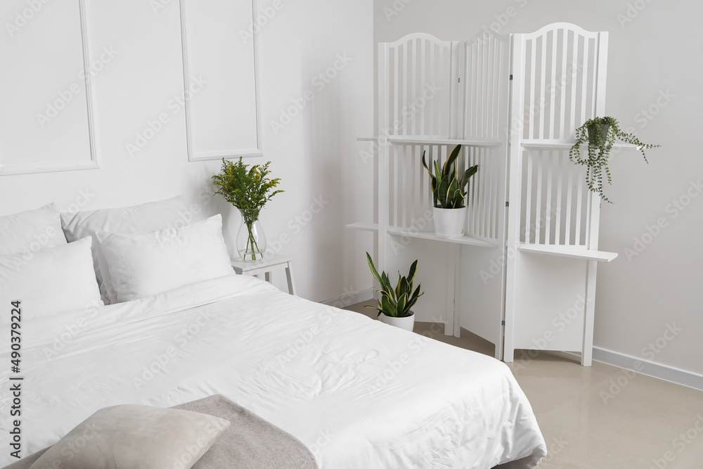 Comfortable bed and folding screen with houseplants near white wall