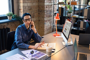 Wall Mural - Serious businesswoman in eyeglasses having a business conversation on mobile phone while working at the table at office with documents and computer