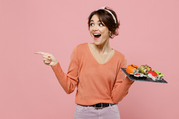 Wall Mural - Young happy fun woman in sweater hold makizushi sushi roll served on black plate traditional japanese food point index finger aside on workspace area mock up isolated on plain pastel pink background