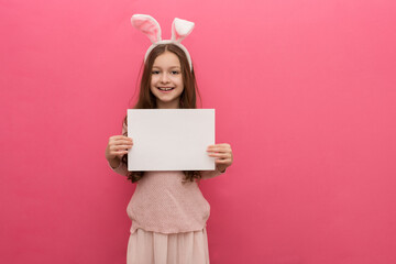 little happy girl with dark hair with hare ears on her head holds a sheet of paper in her hands, space for text, easter card or congratulation, pink background